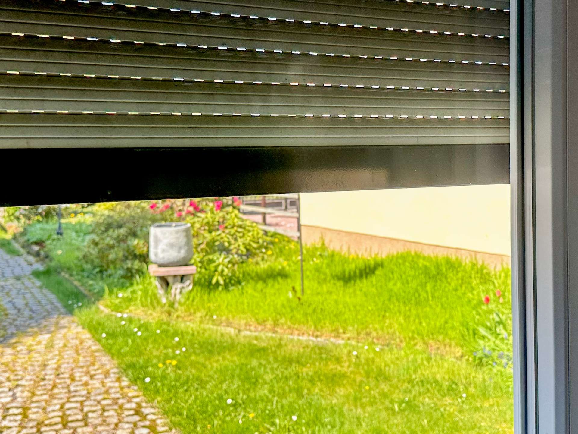 Partially closed metal roller shutter above a window; view of a garden with a stone path, birdbath, and green plants outside.