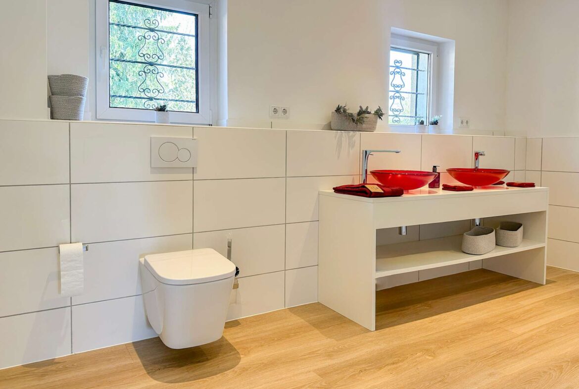 Modern bathroom with two red vessel sinks on a white vanity and open shelving below, finished in white tiles and light wood flooring.