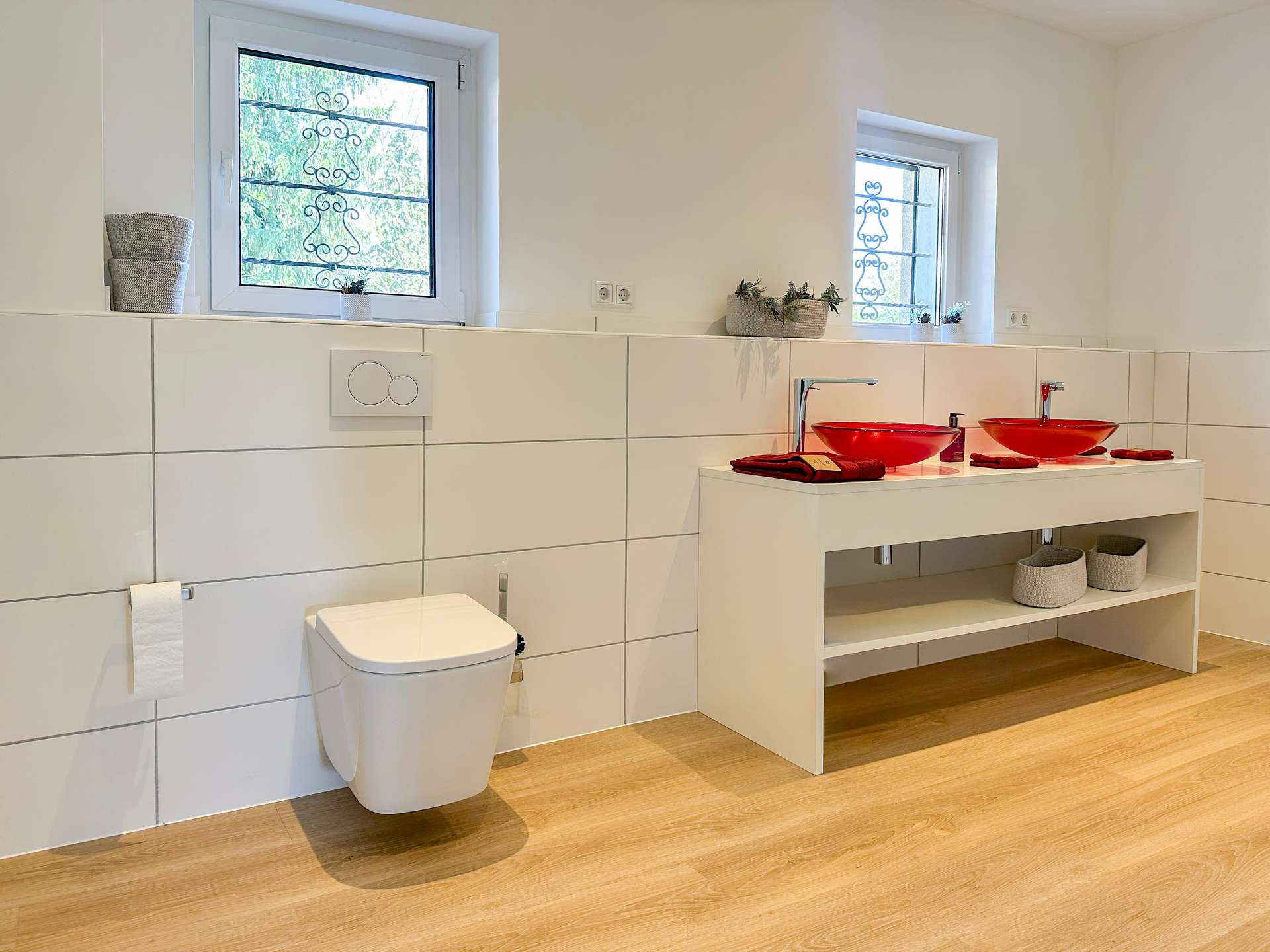 Modern bathroom with two red vessel sinks on a white vanity and open shelving below, finished in white tiles and light wood flooring.