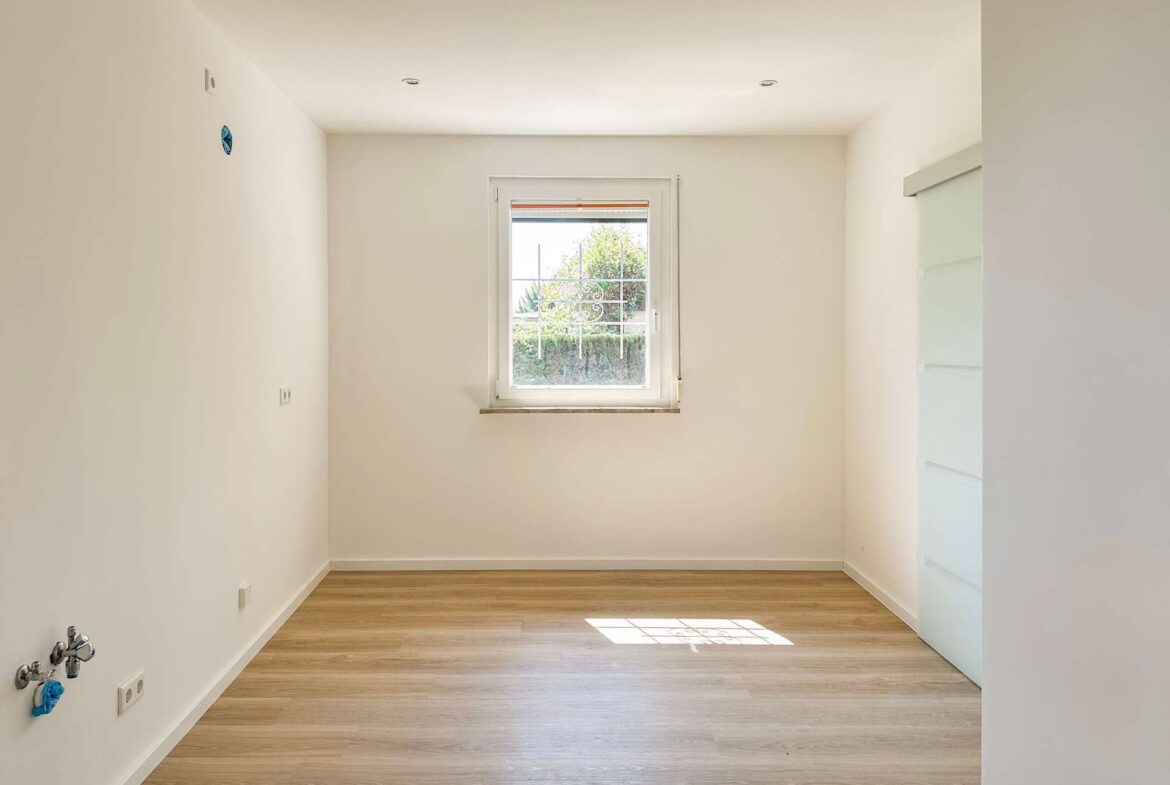 Empty laundry/utility room with exposed water hookups on the left wall, white walls, and a small centered window letting in light.