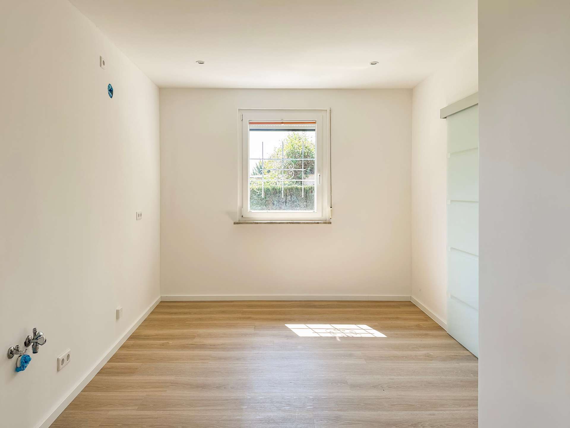 Empty laundry/utility room with exposed water hookups on the left wall, white walls, and a small centered window letting in light.