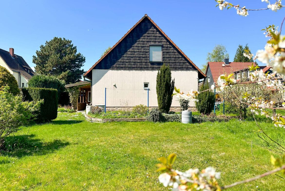 Front yard of a house with a steep triangular roof, white walls, and blooming branches in the foreground on a sunny day.