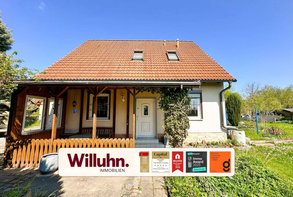 Front view of a house with a red tiled roof, two dormer windows, and a covered porch; a large Willuhn Immobilien sign with award stickers in front.