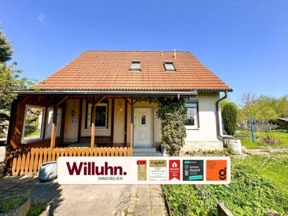 Front view of a house with a red tiled roof, two dormer windows, and a covered porch; a large Willuhn Immobilien sign with award stickers in front.