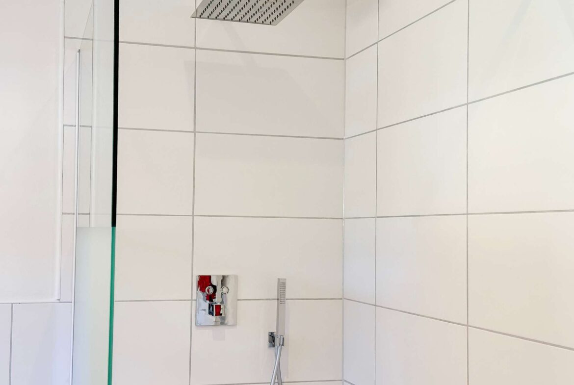Shower area with a large square rain showerhead mounted on tiled wall, glass partition on the left, and a handheld shower hose below.