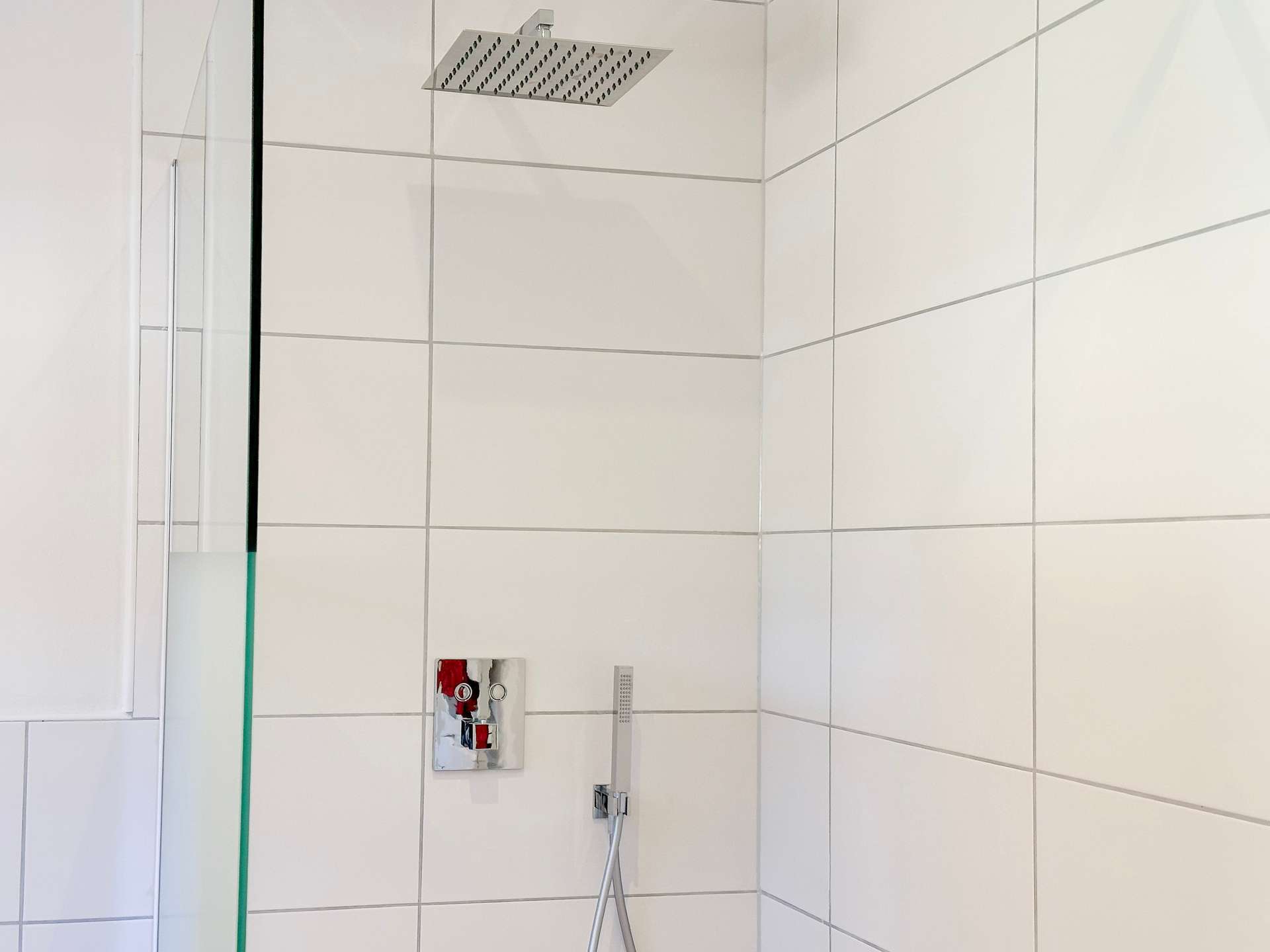 Shower area with a large square rain showerhead mounted on tiled wall, glass partition on the left, and a handheld shower hose below.