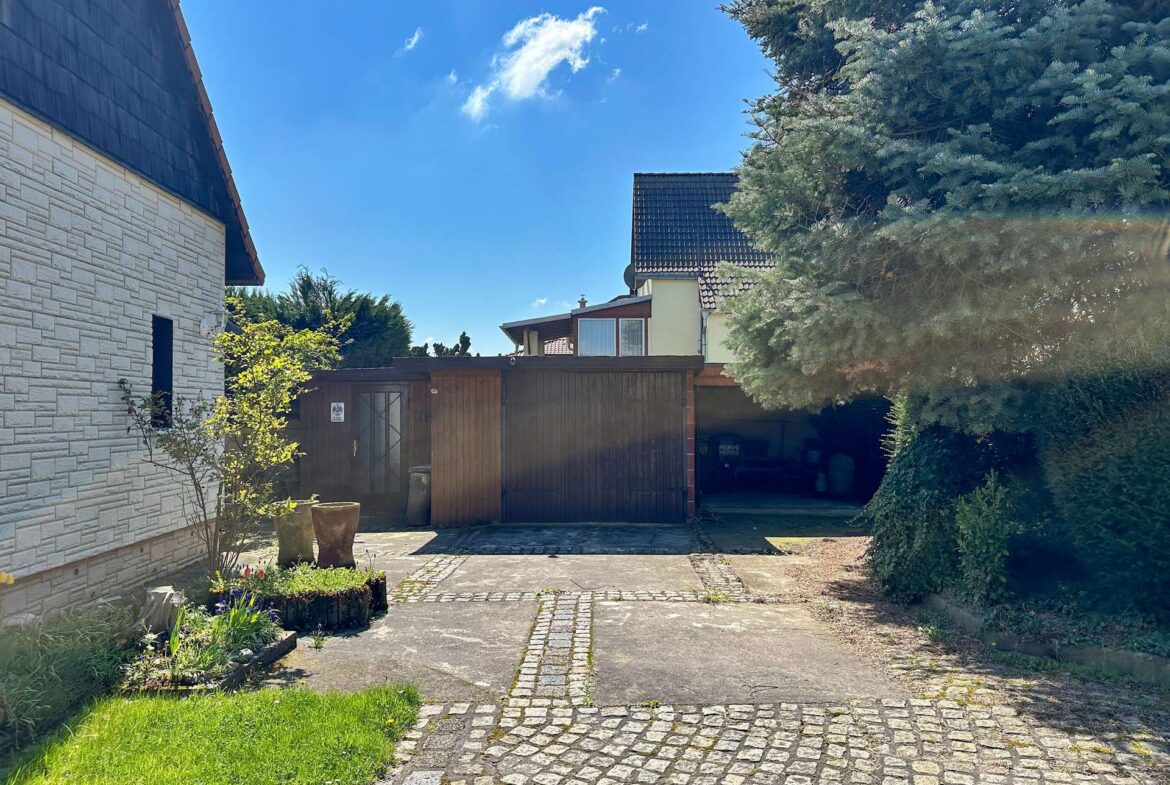 Residential courtyard with a cobblestone path, stone wall on left, wooden gate in the center, and a tall evergreen tree on the right under a blue sky.