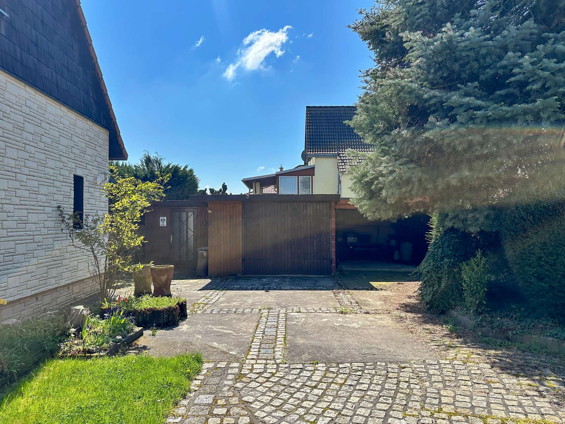 Residential courtyard with a cobblestone path, stone wall on left, wooden gate in the center, and a tall evergreen tree on the right under a blue sky.