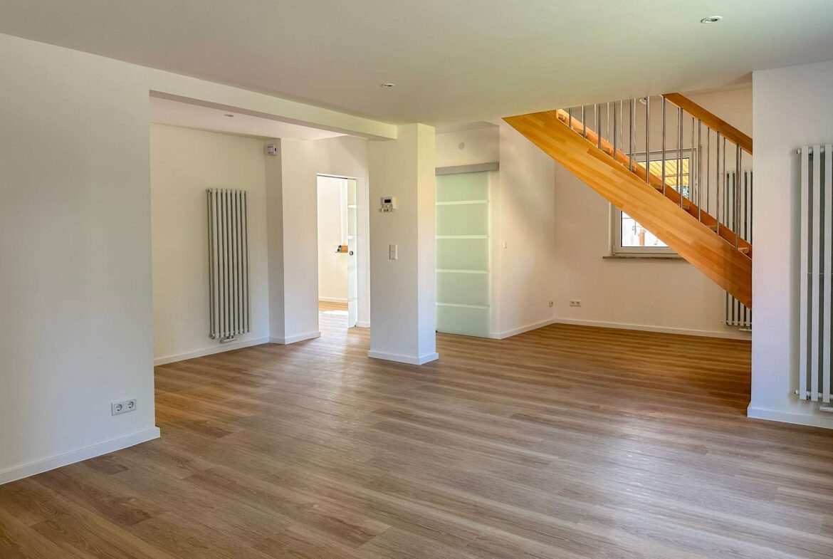 Open, empty living area with light wood floors, white walls, and a wooden staircase with metal balusters on the right side.