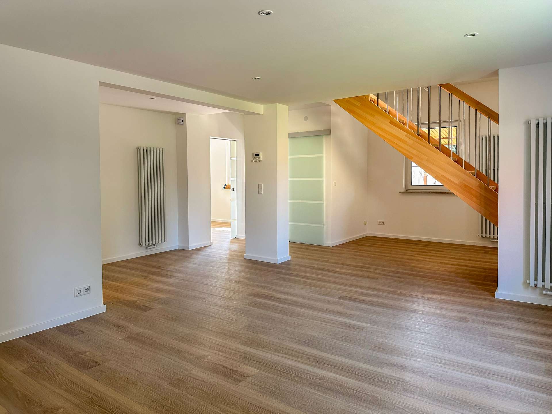 Open, empty living area with light wood floors, white walls, and a wooden staircase with metal balusters on the right side.