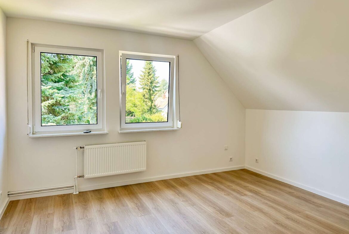 Bright empty room with two side-by-side windows, radiator, and light wood floor under a sloped ceiling.