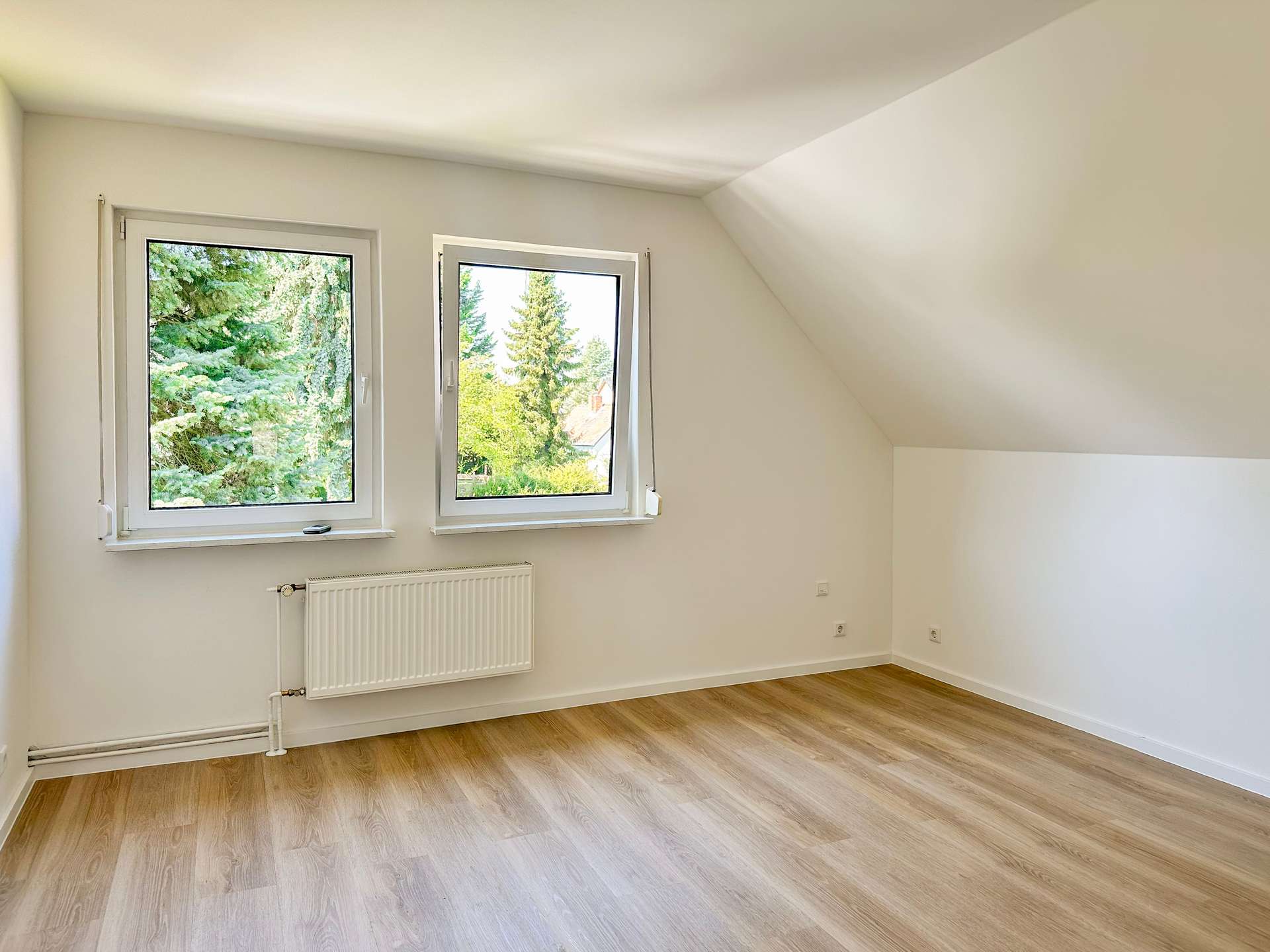 Bright empty room with two side-by-side windows, radiator, and light wood floor under a sloped ceiling.
