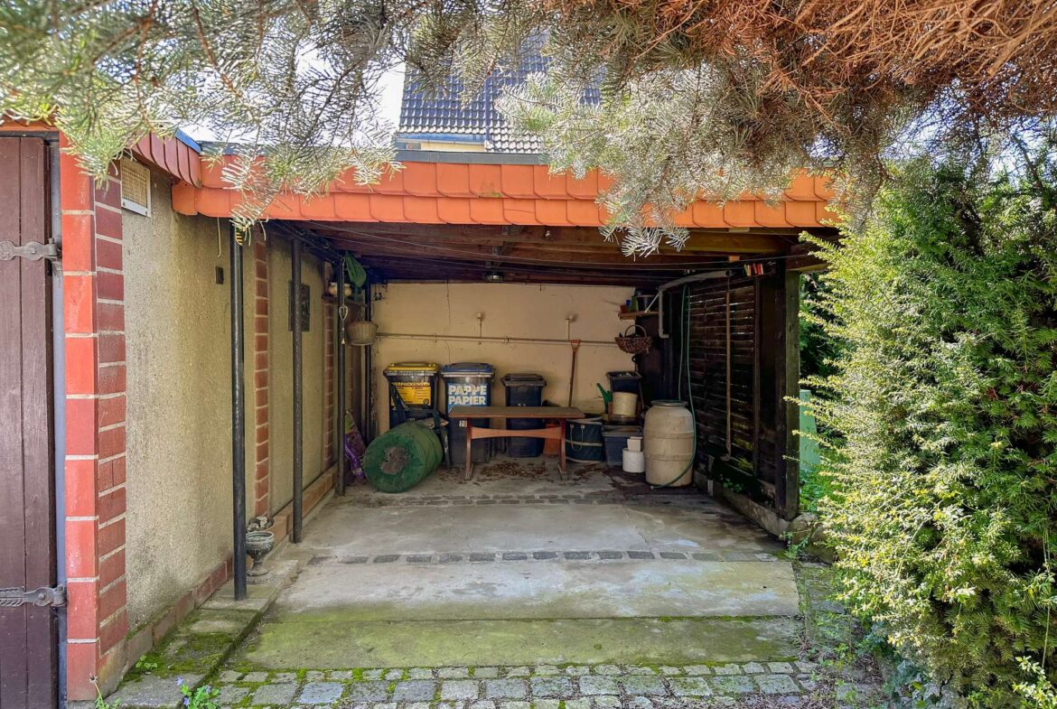 Open carport under a red-tiled canopy, cluttered with bins, shelves, and a wooden table on a mossy concrete floor.