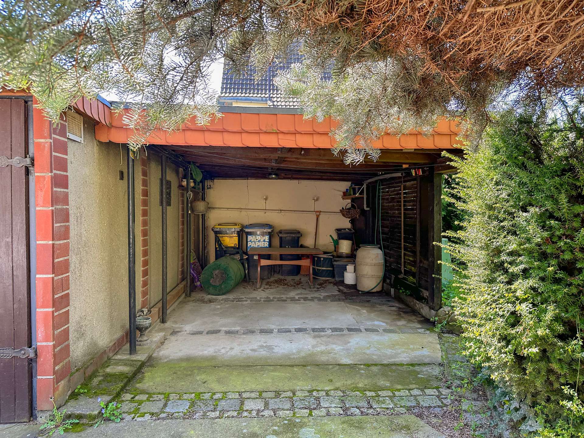 Open carport under a red-tiled canopy, cluttered with bins, shelves, and a wooden table on a mossy concrete floor.