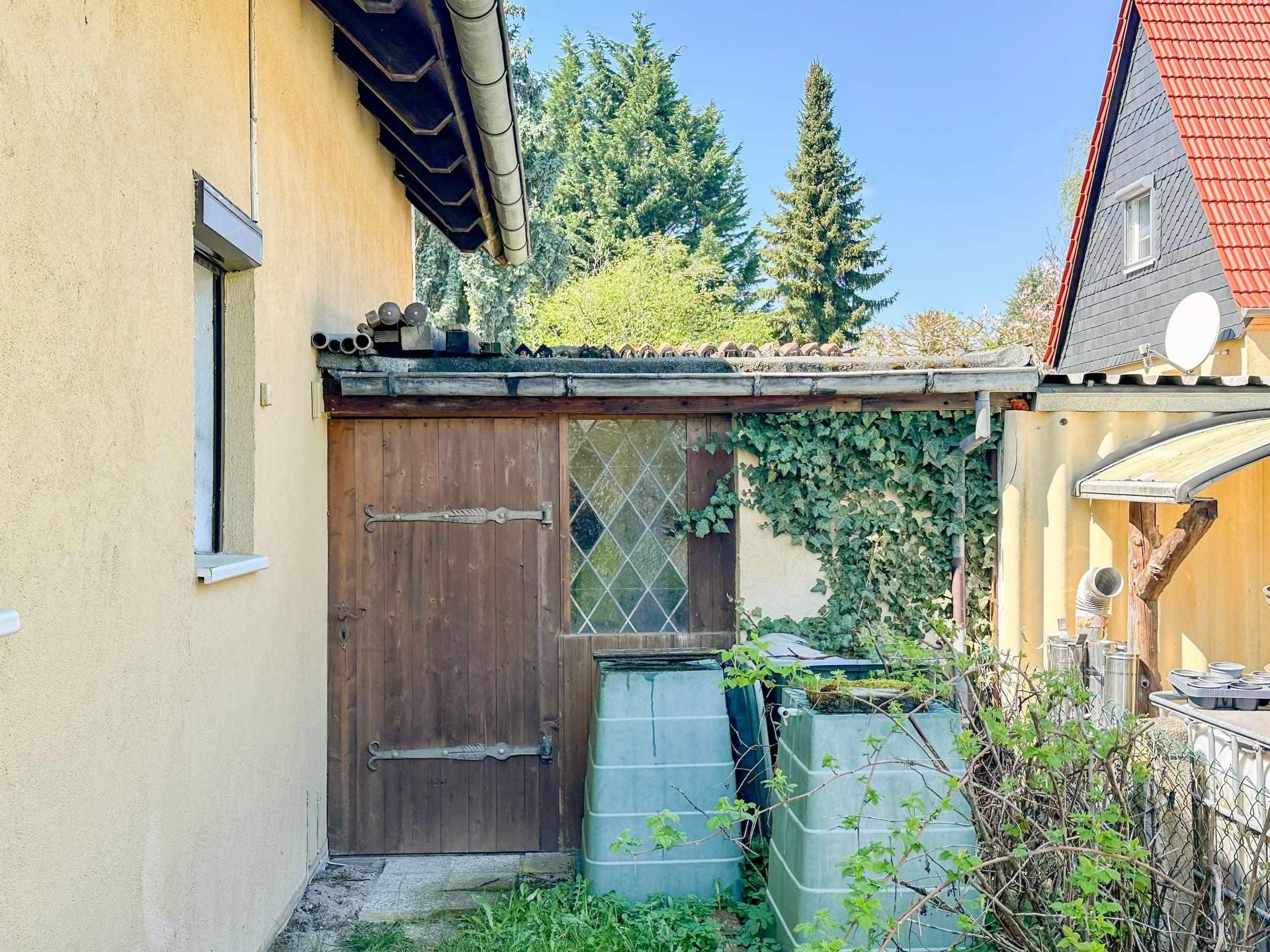 Narrow side yard between houses with rain barrels, ivy, and a wooden gate against a blue sky.