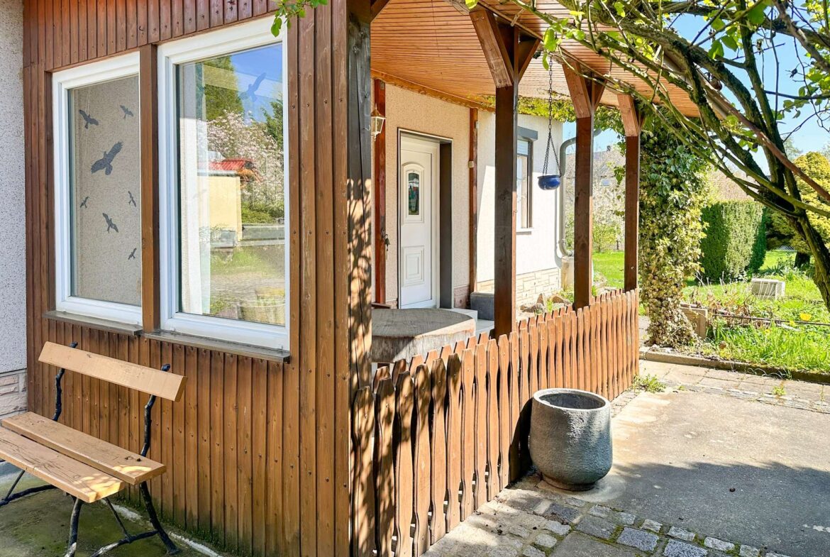 Wood-paneled house with a small porch, two white-framed windows, and a wooden picket fence; a bench sits along the wall.