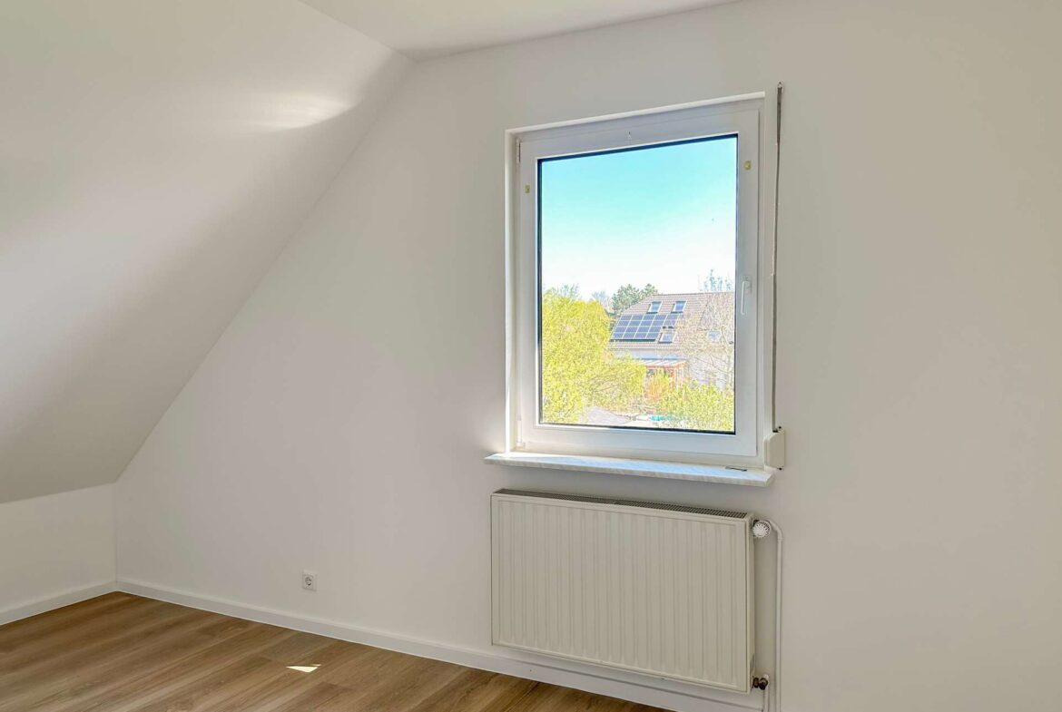 Bright empty attic room with a single window, sloped left wall, and wood-look floor; radiator under the window.