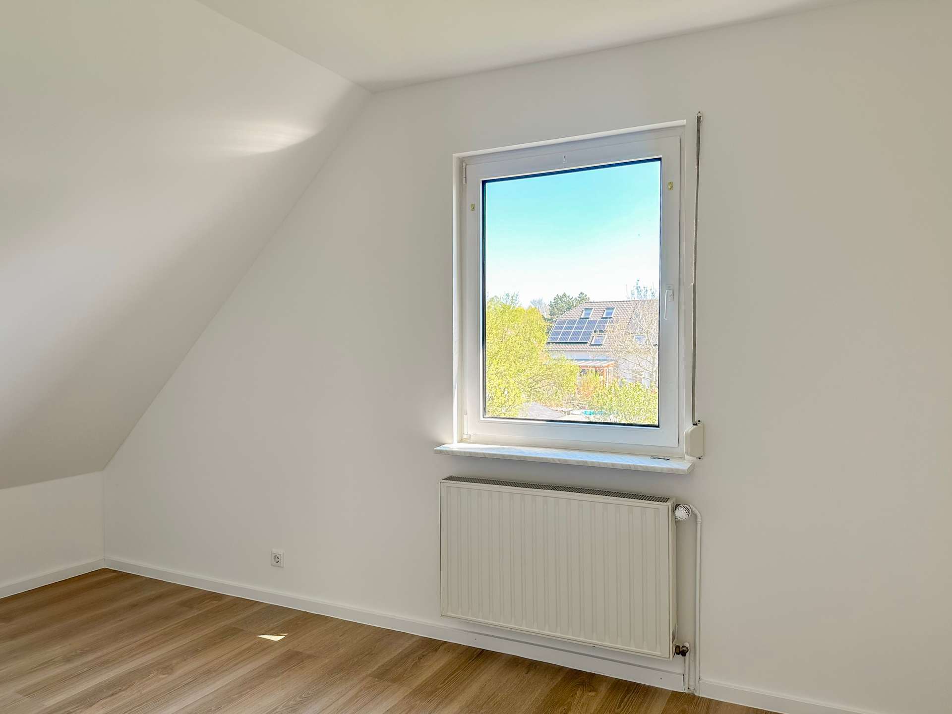 Bright empty attic room with a single window, sloped left wall, and wood-look floor; radiator under the window.