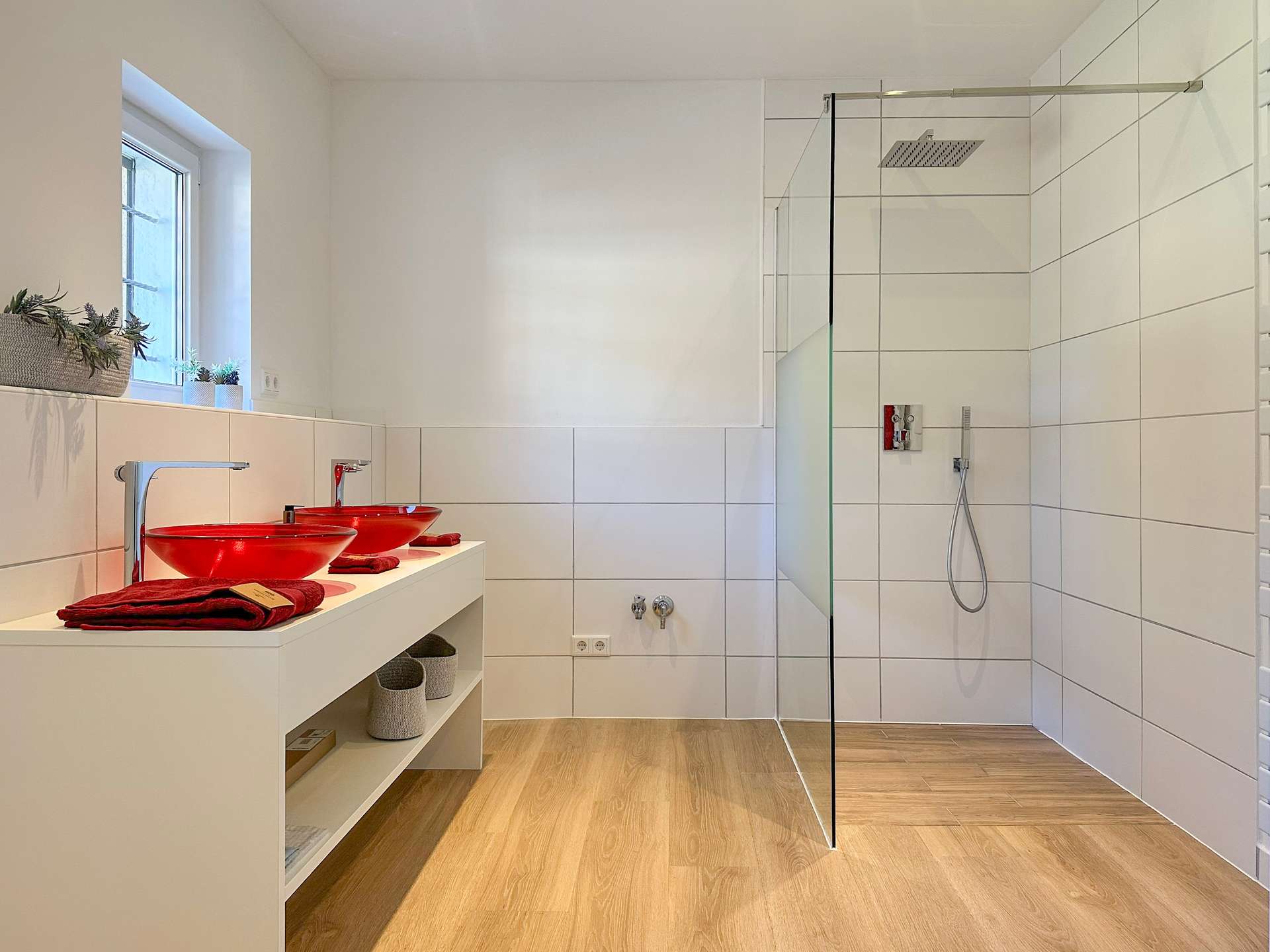 Modern white bathroom with a double red vessel sink on a white vanity, window with potted plants, and a glass shower enclosure.