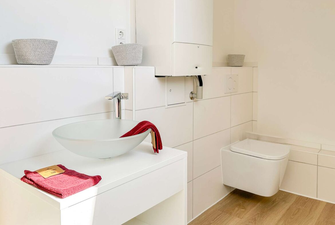 Modern white bathroom with a wall-mounted sink and red towels, plus wicker baskets and a toilet to the right.