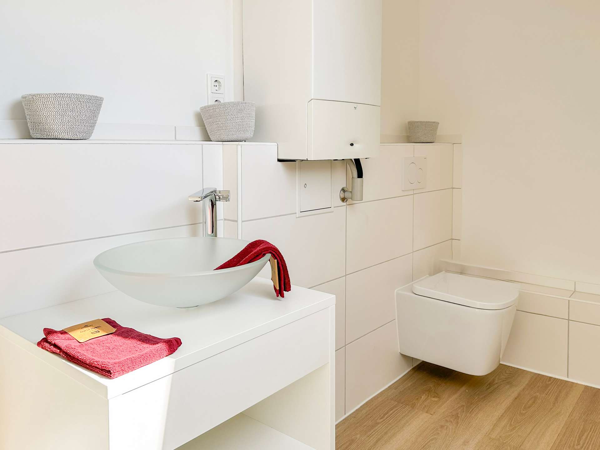 Modern white bathroom with a wall-mounted sink and red towels, plus wicker baskets and a toilet to the right.