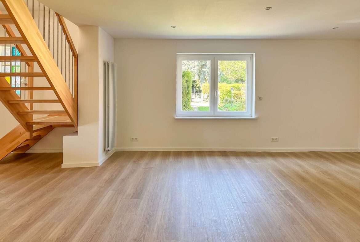 Empty living room with light wood flooring, white walls, and a wooden staircase on the left; view of a garden through a central window