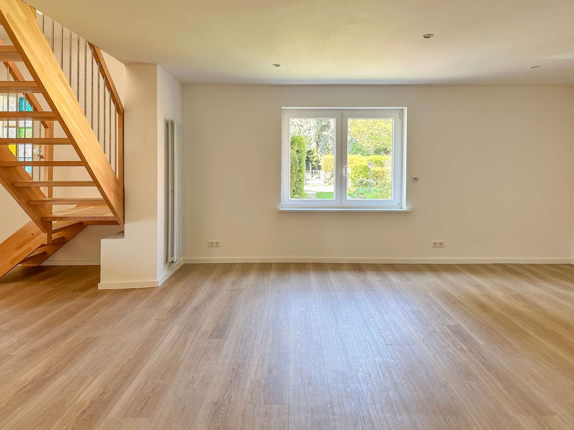 Empty living room with light wood flooring, white walls, and a wooden staircase on the left; view of a garden through a central window