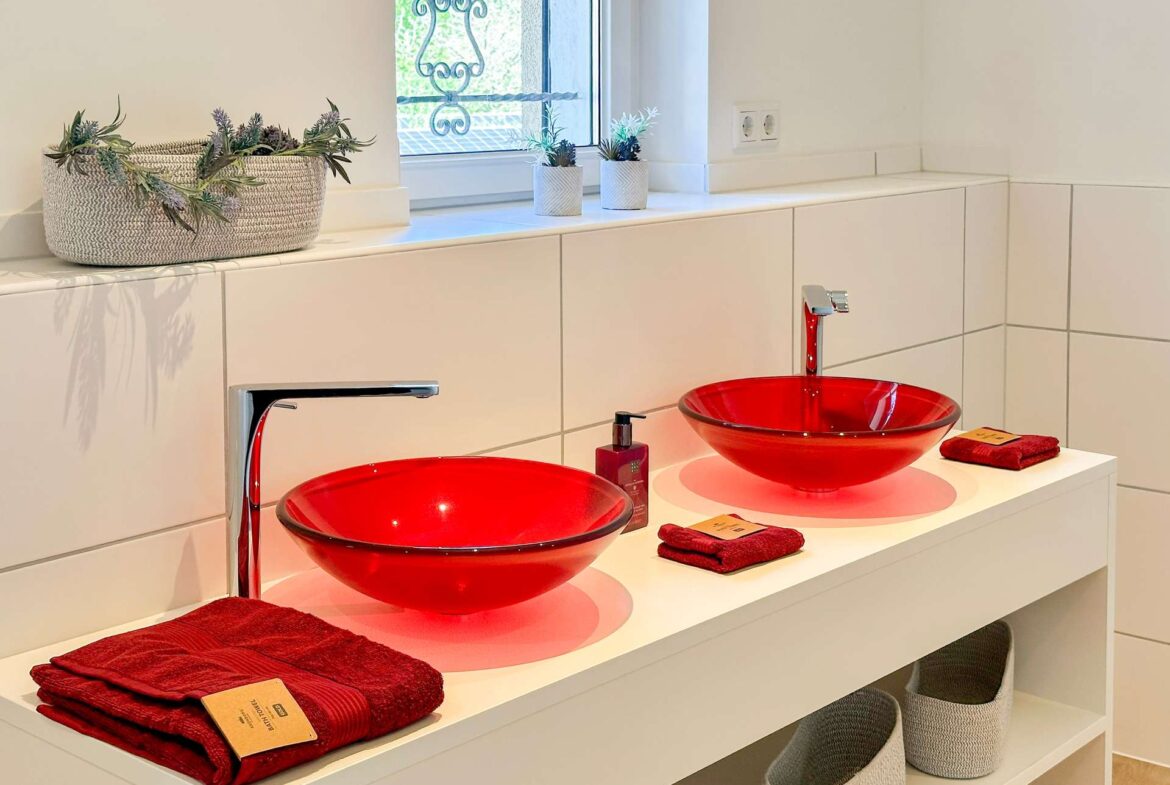 Double vanity with two red glass sinks, white countertop, red towels, and small potted plants by a window in a bright bathroom.