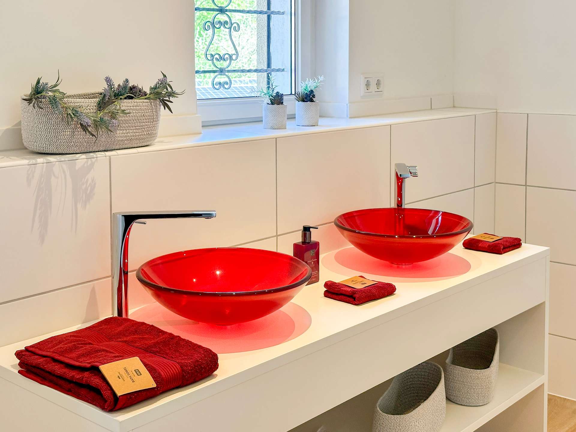 Double vanity with two red glass sinks, white countertop, red towels, and small potted plants by a window in a bright bathroom.