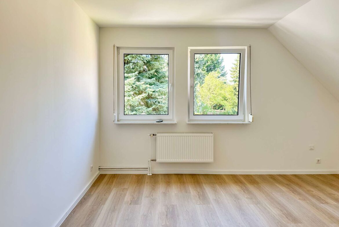 Small empty room with two side-by-side windows showing green trees, radiator below, and light wood flooring.