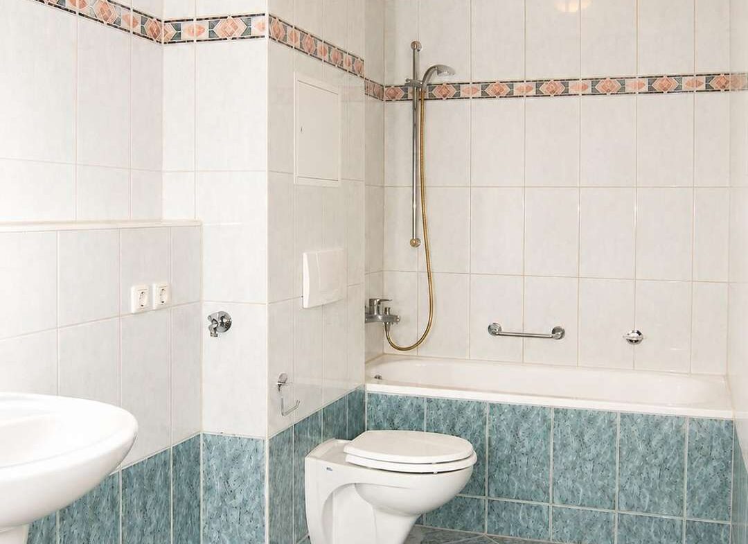 Bright bathroom with white wall tiles, teal lower wall and floor tiles, wall-mounted toilet, and a bathtub with a handheld shower.