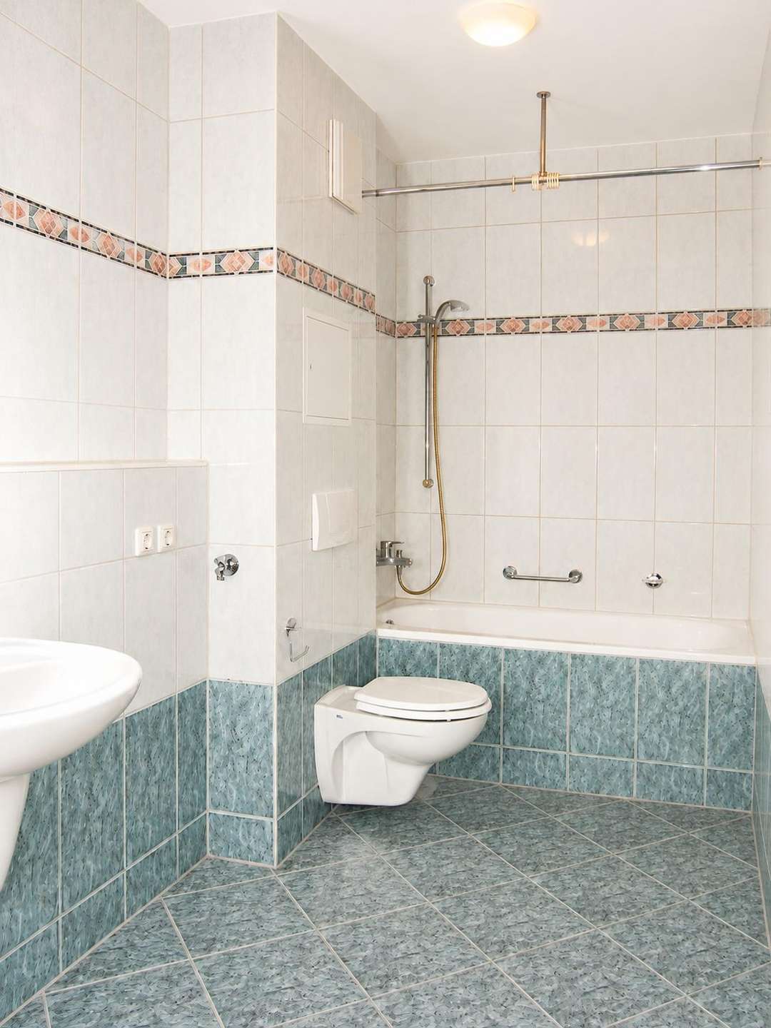 Bright bathroom with white wall tiles, teal lower wall and floor tiles, wall-mounted toilet, and a bathtub with a handheld shower.