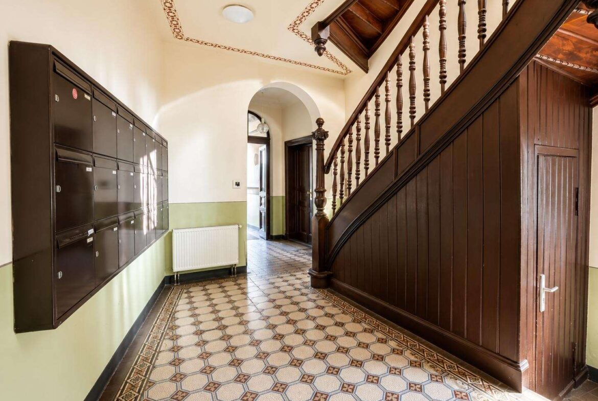 Interior hallway with a dark wooden staircase on the right, row of black mailboxes on the left, and a tiled patterned floor with pale green wainscoting under the radiator.