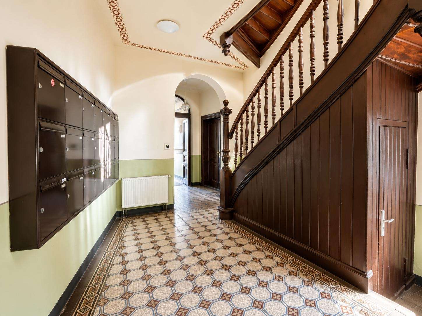 Interior hallway with a dark wooden staircase on the right, row of black mailboxes on the left, and a tiled patterned floor with pale green wainscoting under the radiator.