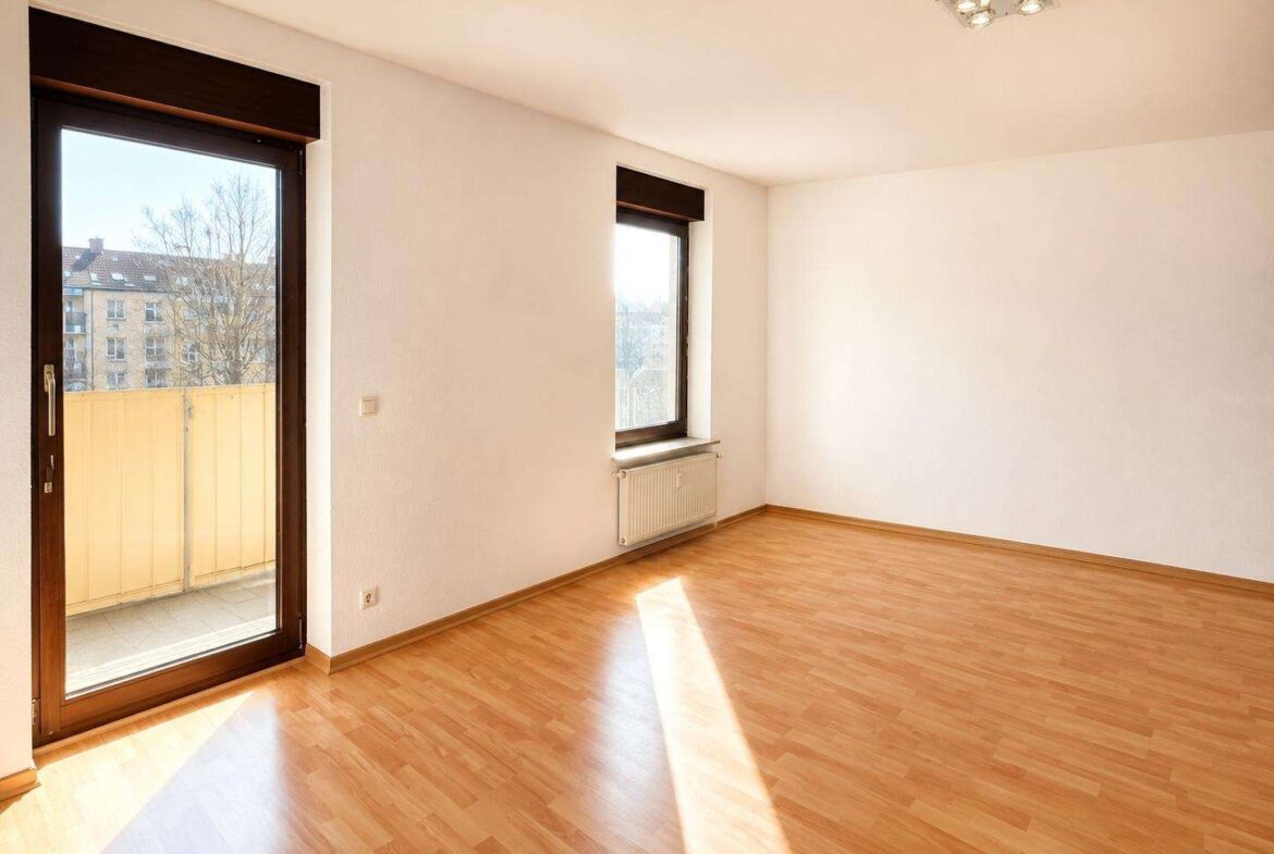 Bright, empty living room with light wood flooring, white walls, a large window, and a balcony door letting in sunlight from outside.