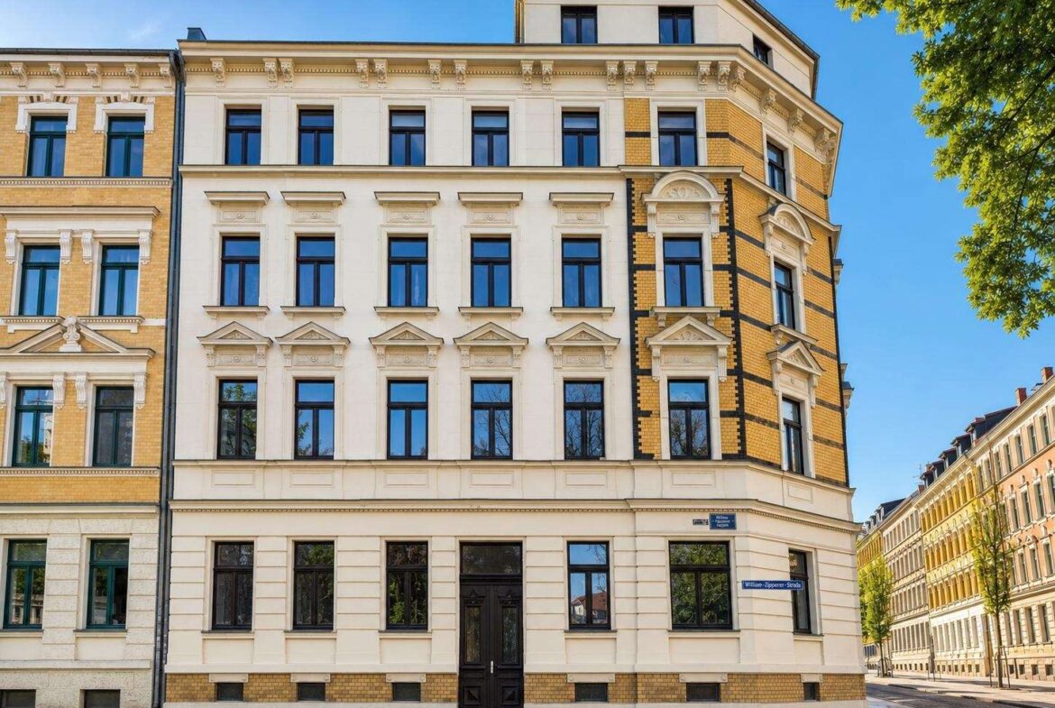 Historic four-story building with a white facade and ornate window surrounds on a sunny city street, flanked by beige brick buildings and green trees against a blue sky.