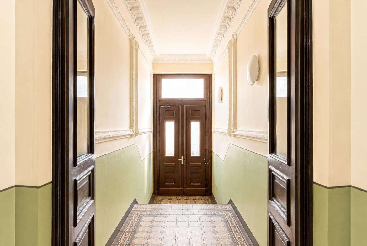 Narrow vintage hallway with ornate plaster crown molding, beige walls, green wainscoting, and dark wood doors at the far end.