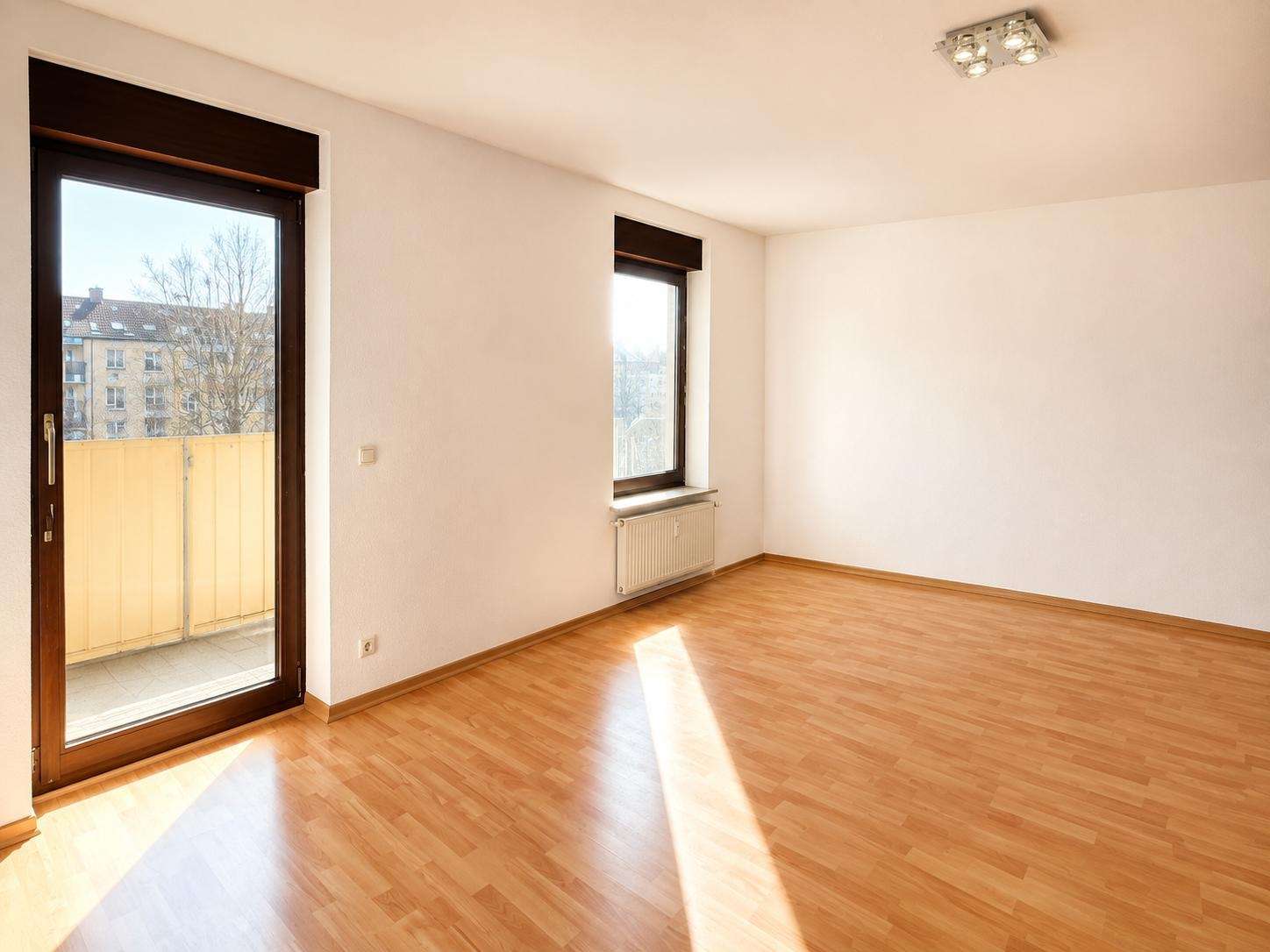 Sunlit empty room with light wood floor, white walls, a glass balcony door on the left and a window with a radiator beneath.