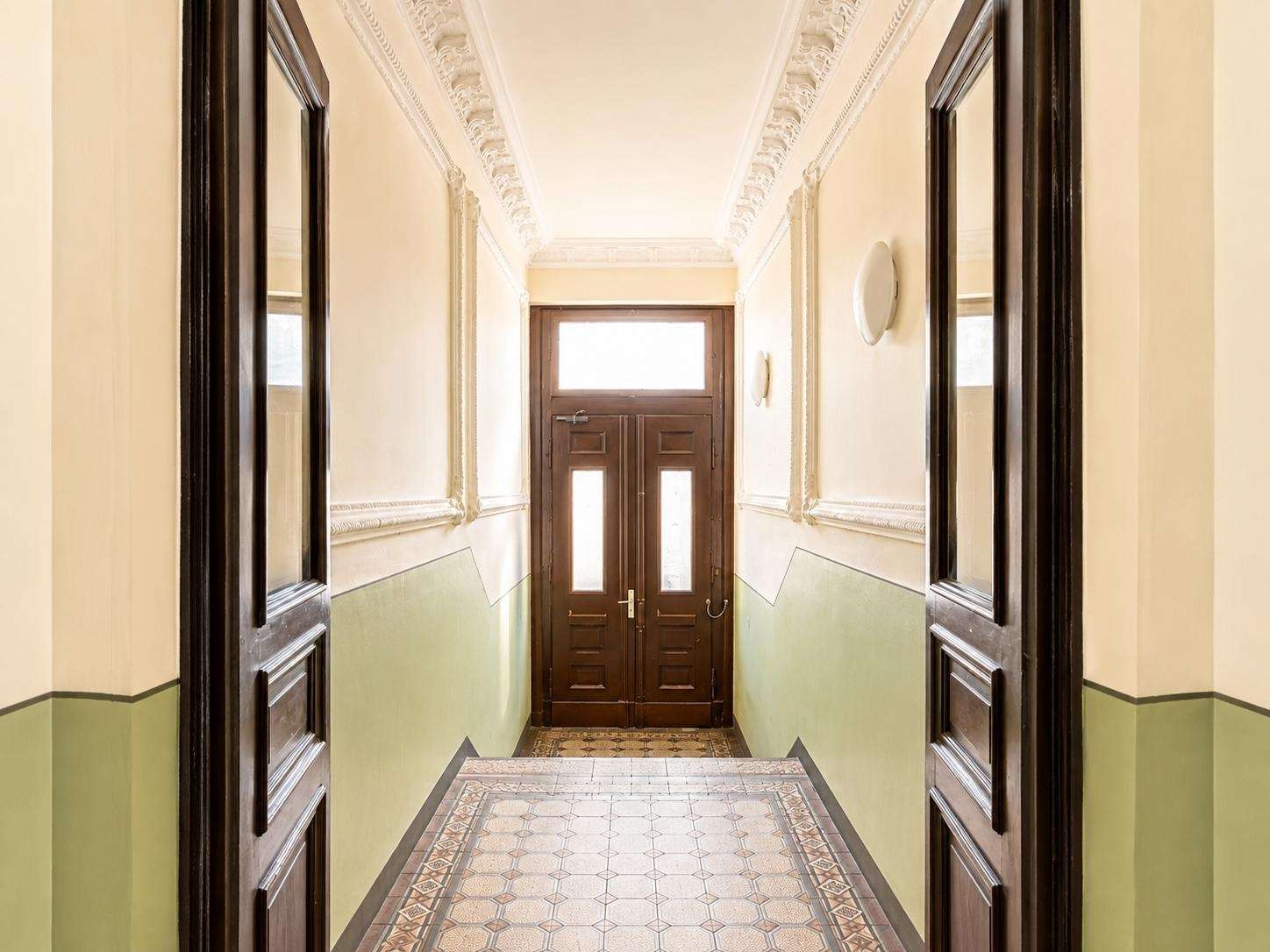 Narrow hallway with cream walls, decorative ceiling molding, and green wainscoted lower walls leading to dark wooden double doors at the end.