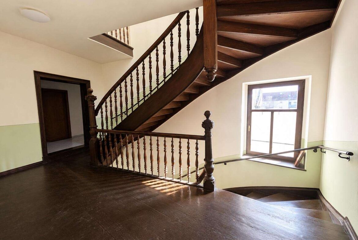 Curved wooden staircase with ornate balusters in a bright stairwell, sunlight from a large window on the right.