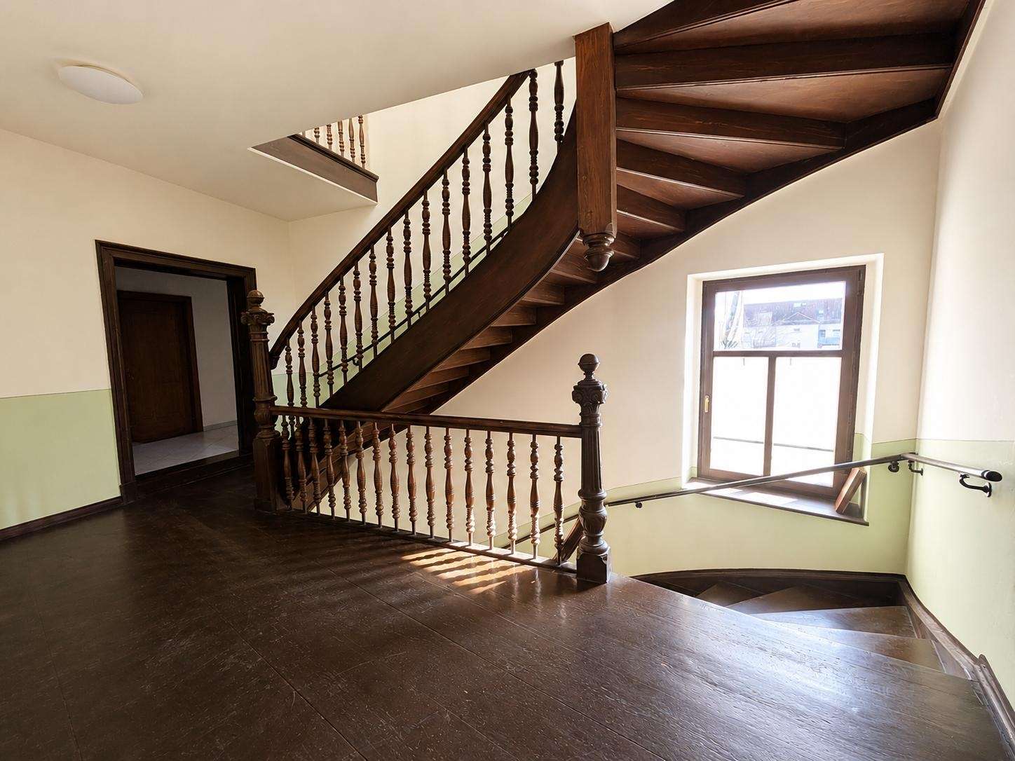 Curved wooden staircase with ornate balusters in a bright stairwell, sunlight from a large window on the right.