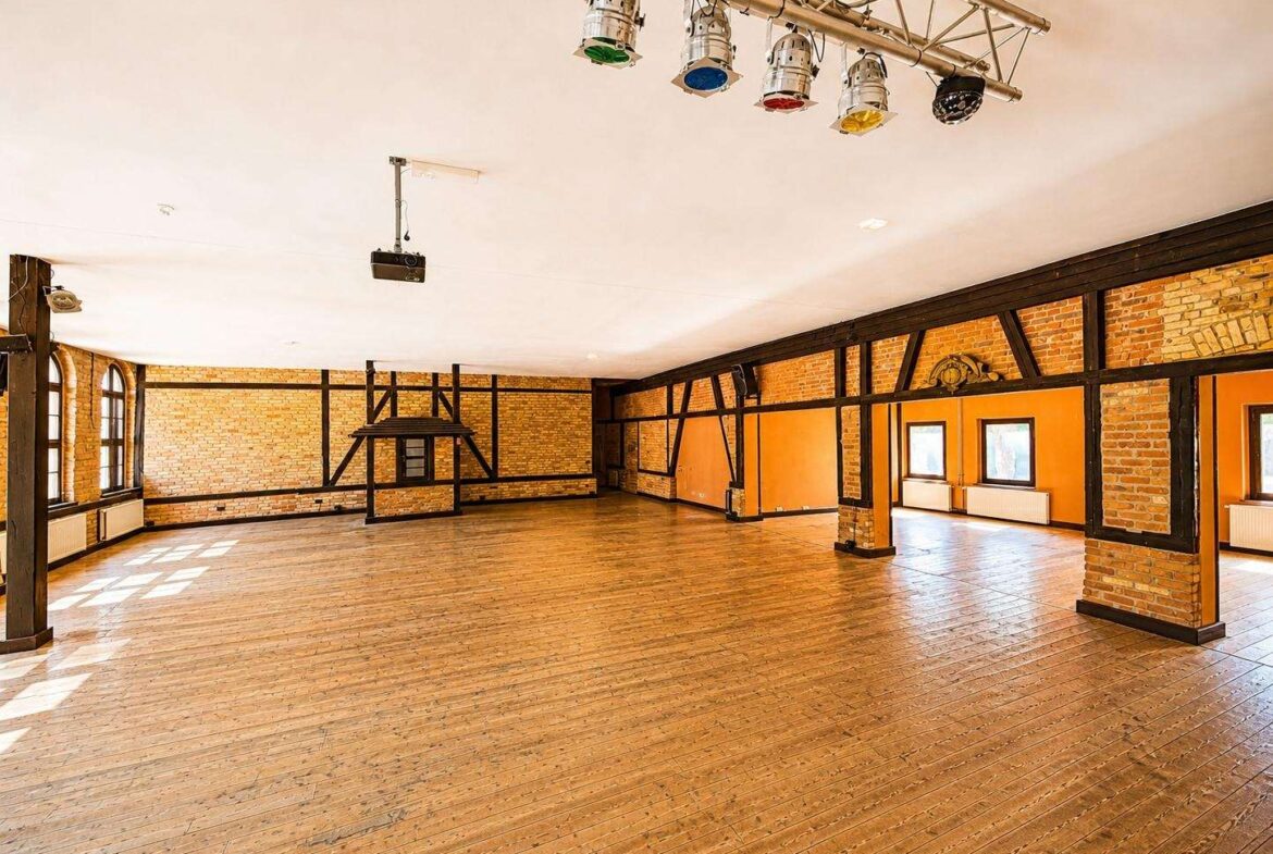 Spacious brick-walled event hall with a wooden floor, black timber beams, a raised stage frame, and hanging stage lights above.