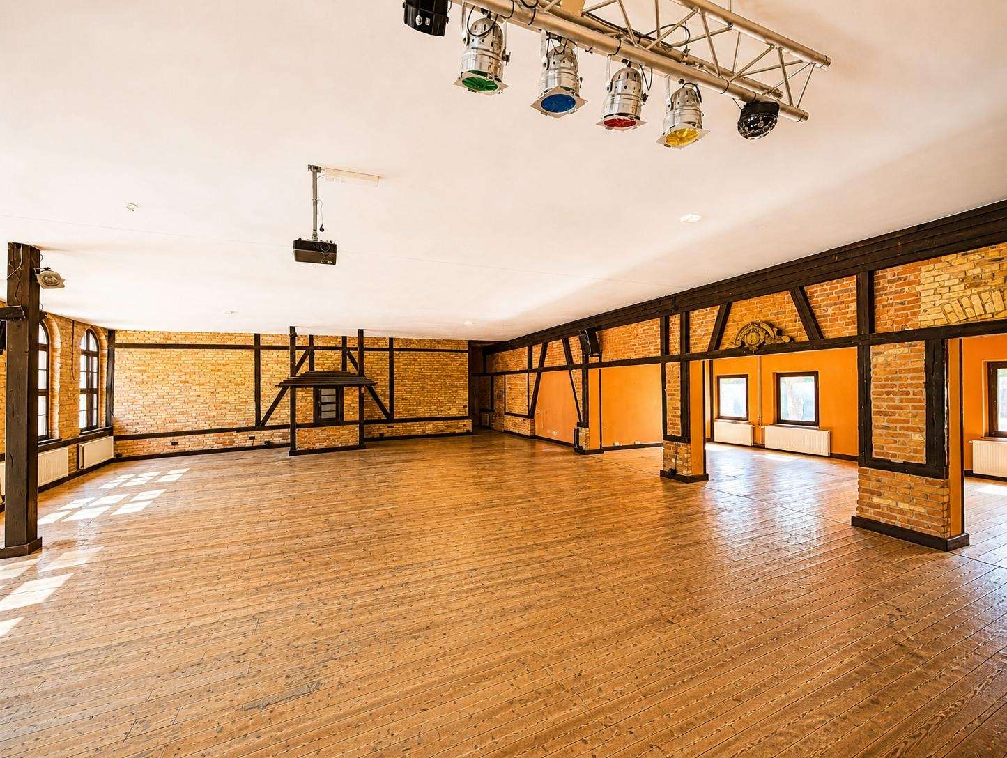 Spacious brick-walled event hall with a wooden floor, black timber beams, a raised stage frame, and hanging stage lights above.