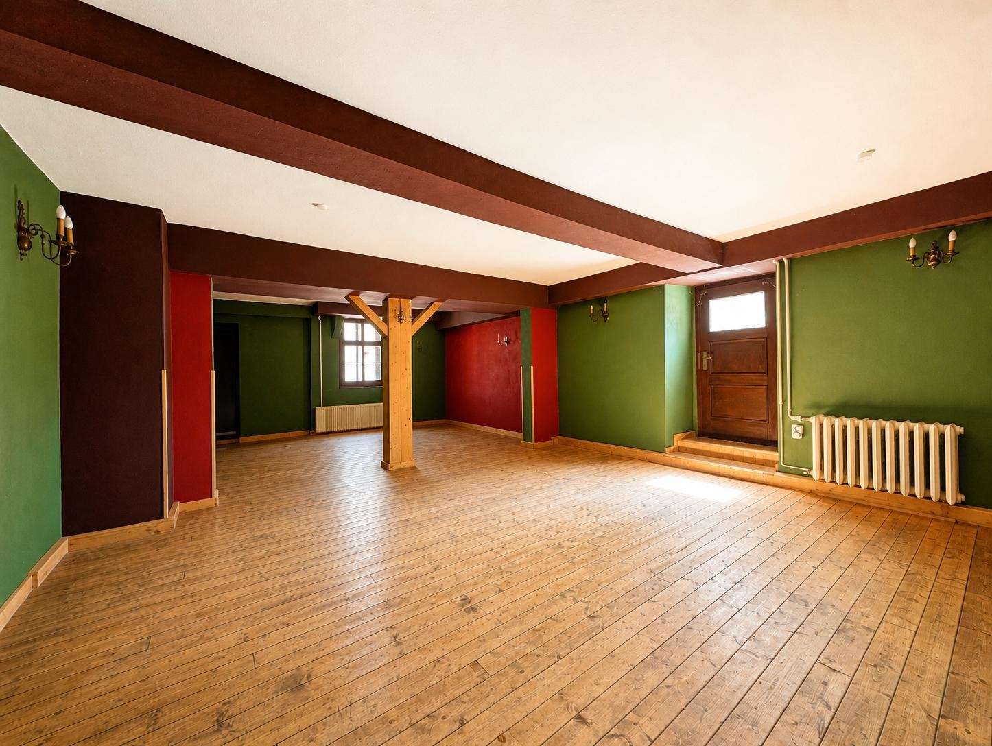 Spacious empty room with green walls, red accents, wooden floor and ceiling beams; central wooden post and radiator by the door.