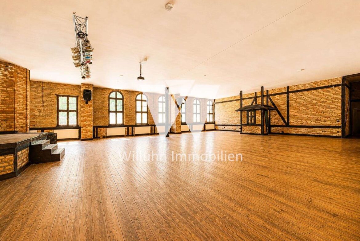 Spacious brick-walled event hall with a wooden floor, arched windows, and an empty stage on the left; overhead lighting rig is visible above.