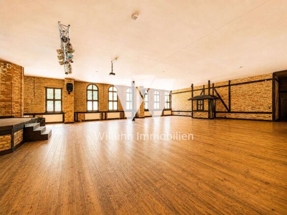 Spacious brick-walled event hall with a wooden floor, arched windows, and an empty stage on the left; overhead lighting rig is visible above.