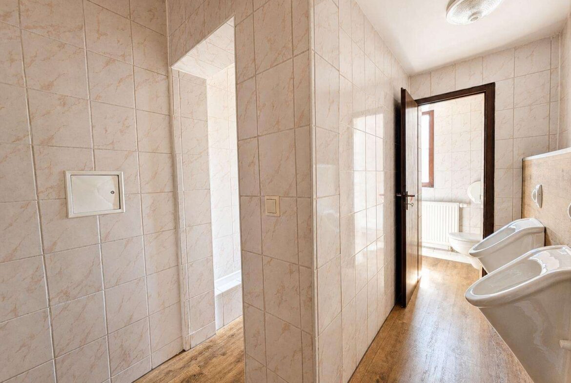 Public bathroom with tiled walls, sinks along the right, and an open wooden door revealing a toilet area beyond a radiator.