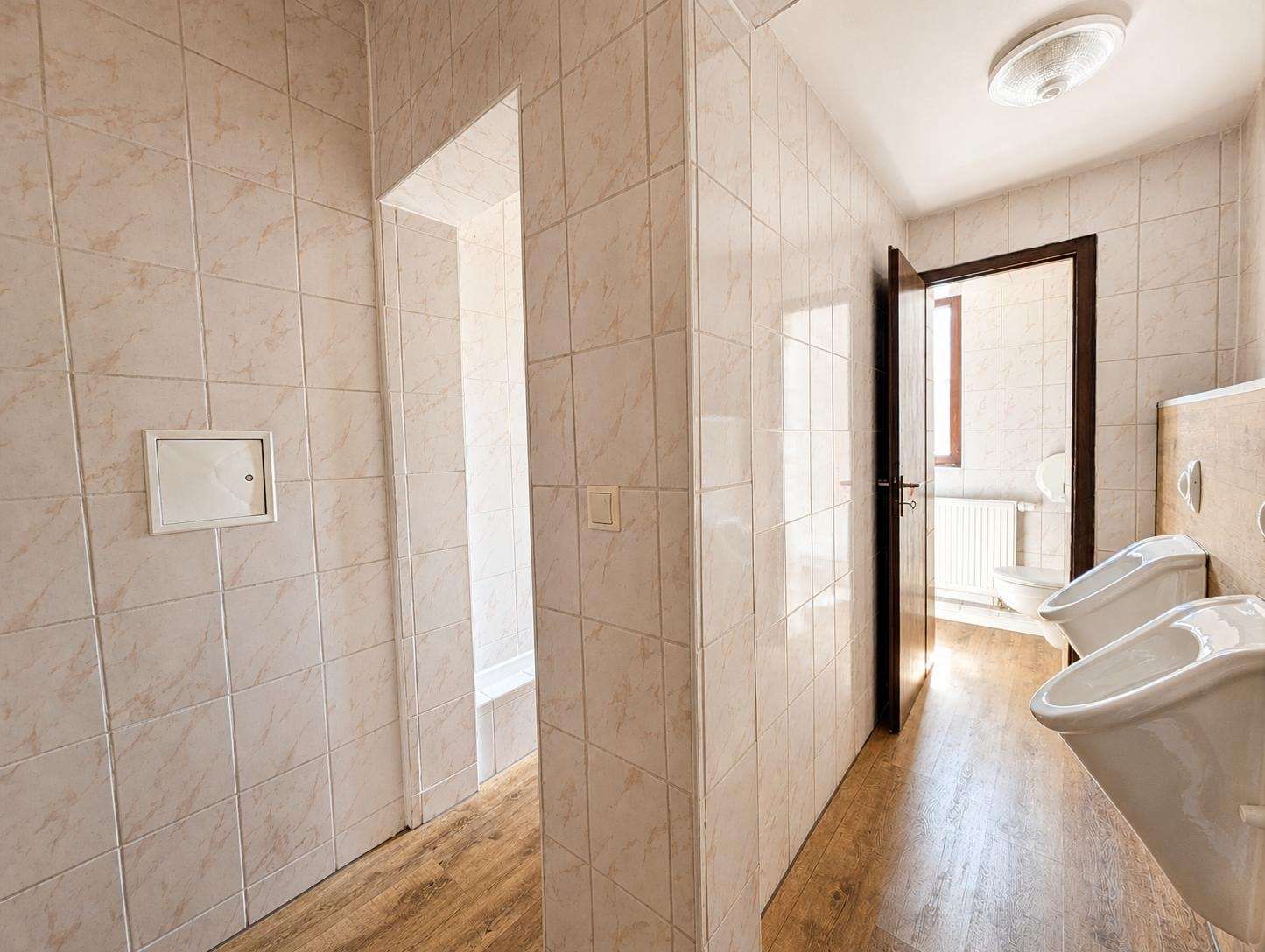 Public bathroom with tiled walls, sinks along the right, and an open wooden door revealing a toilet area beyond a radiator.