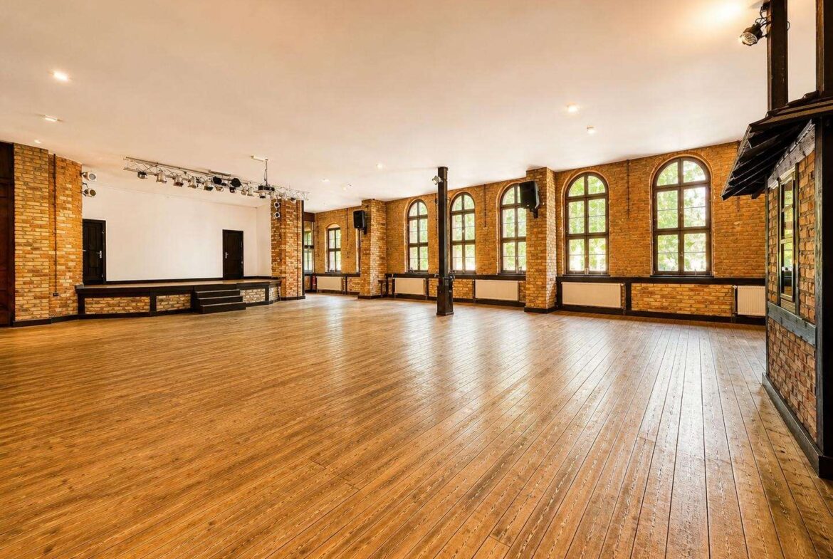 Spacious brick-walled event hall with arched windows, wooden floor, and a small stage on the left.