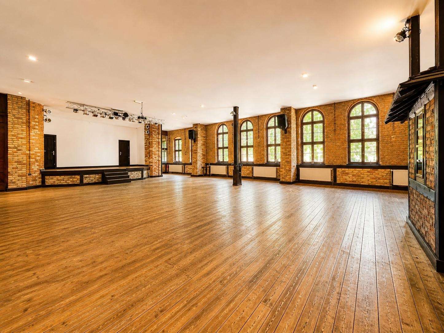 Spacious brick-walled event hall with arched windows, wooden floor, and a small stage on the left.
